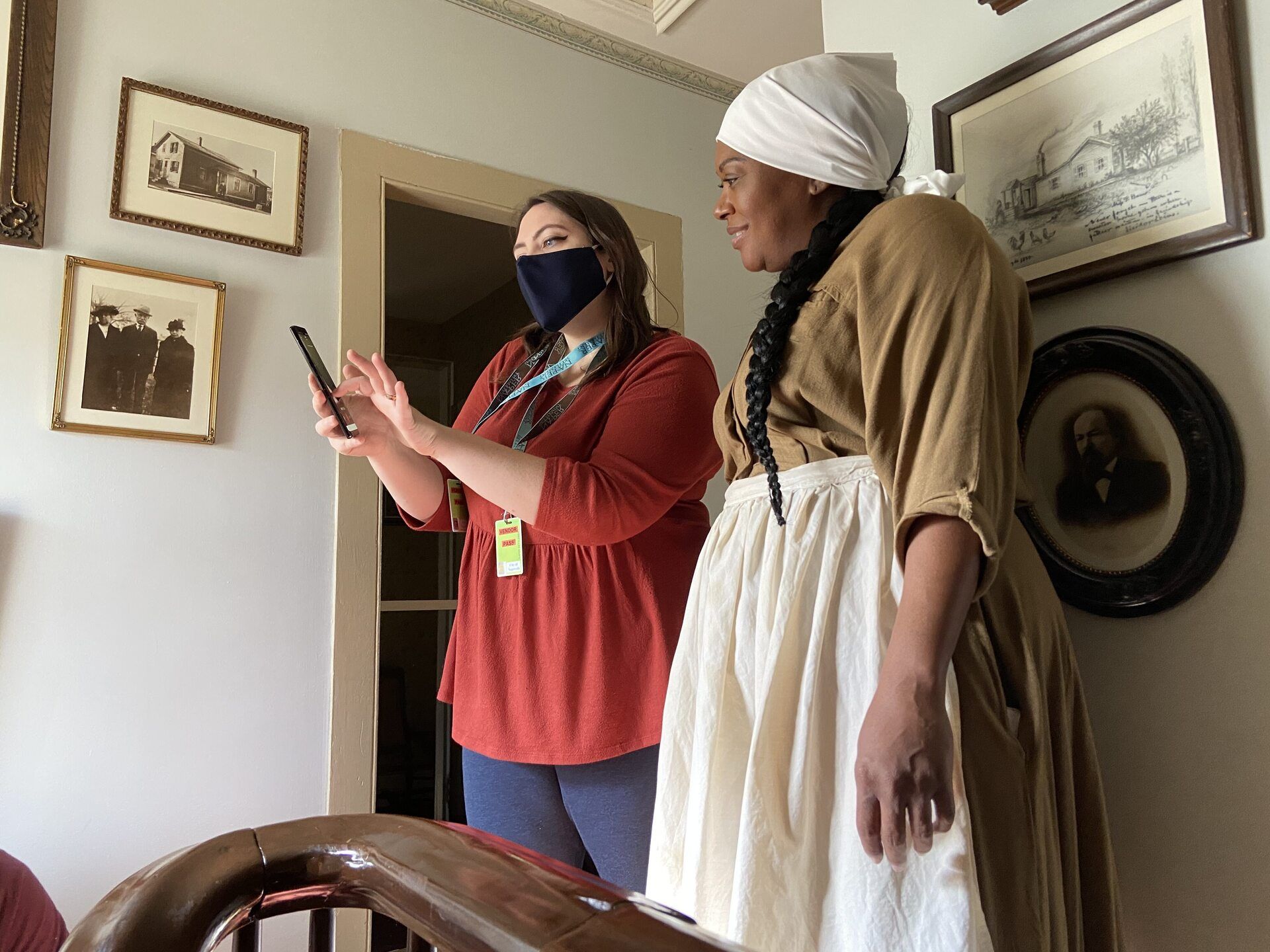 A photo of Valentina from Night Kitchen and an actress behind the scenes of a shoot. Valentina is wearing a face mask and holding up a phone. The actress is wearing a Freedom Seeker costume with an apron and head cloth.